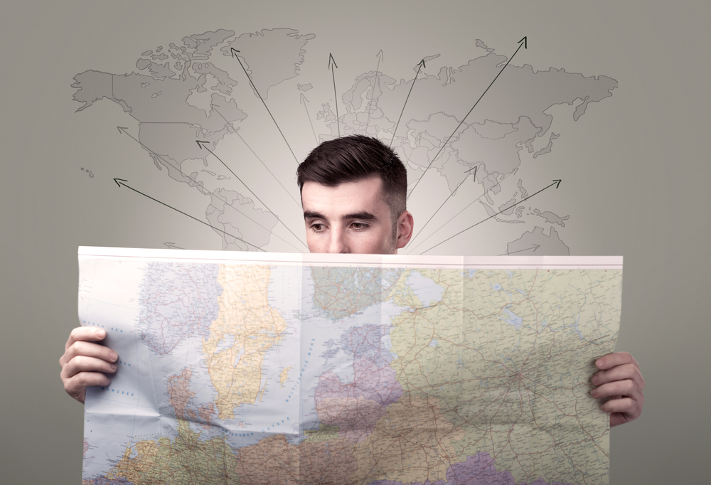 Handsome young man holding a map with world map and arrows behind him